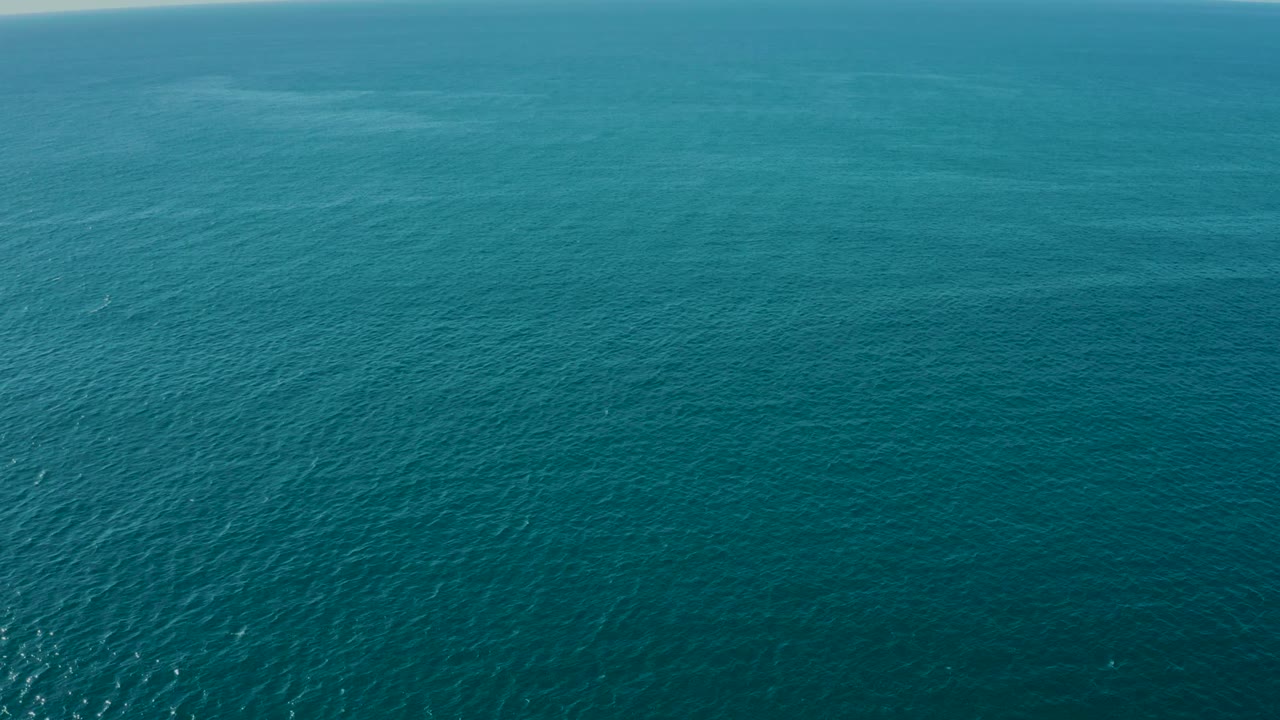 Aerial frame over La Fortuna coastal water with the sun glint reduced from the earlier frames, fine wave texture across the blue surface, a faint horizon, and no visible people.