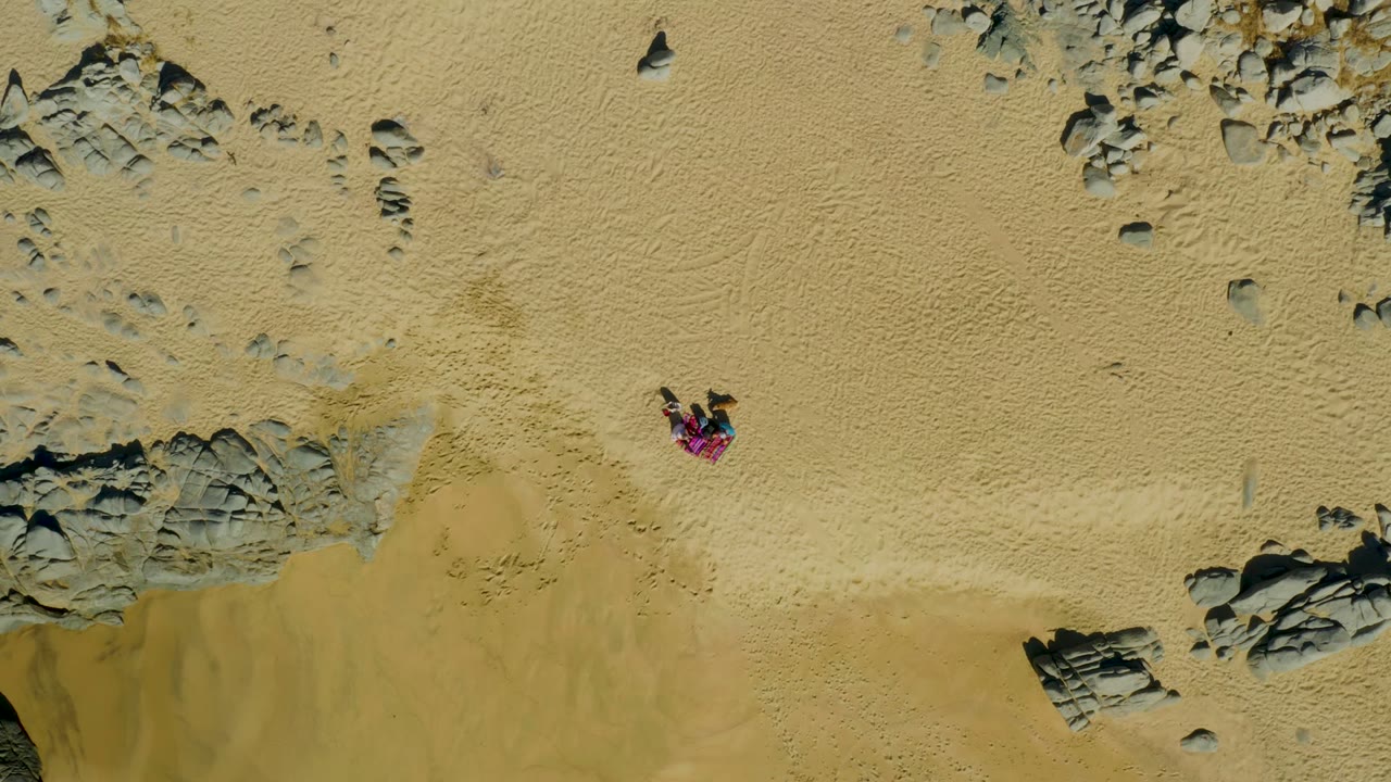 Overhead view in La Fortuna, Baja California Sur, showing a small person in a pink top on pale sand near scattered gray rock clusters.