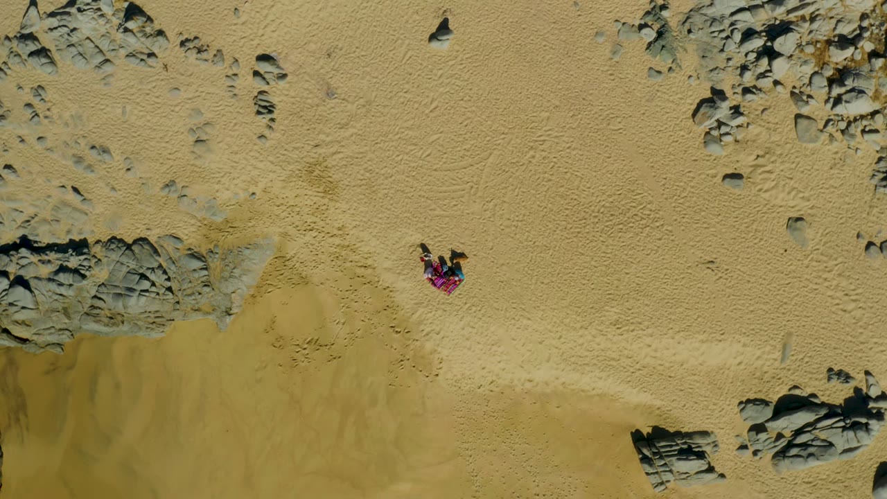 La Fortuna overhead beach view with the person in pink near the upper sand field and dark rock patches framing the sandy terrain.
