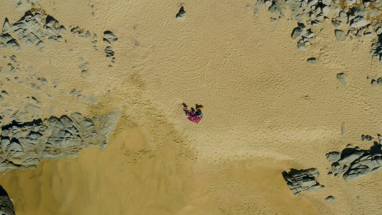 Overhead view in La Fortuna, Baja California Sur, Mexico, showing pale sand, scattered rock formations, wet sand at the lower edge, and a person in a pink top lying near the center-left.