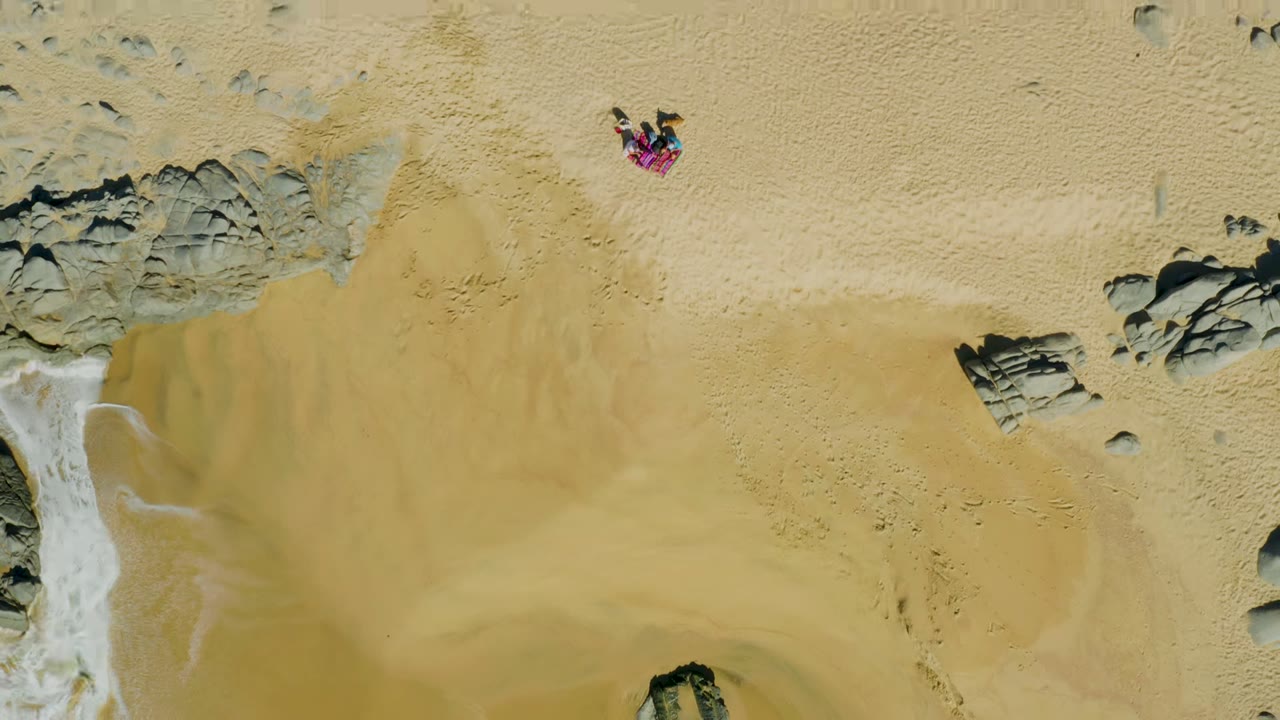 Overhead view at La Fortuna, East Cape, Baja California Sur showing one person on a bright towel above wet sand, with gray rocks and shallow wash along the lower left edge.