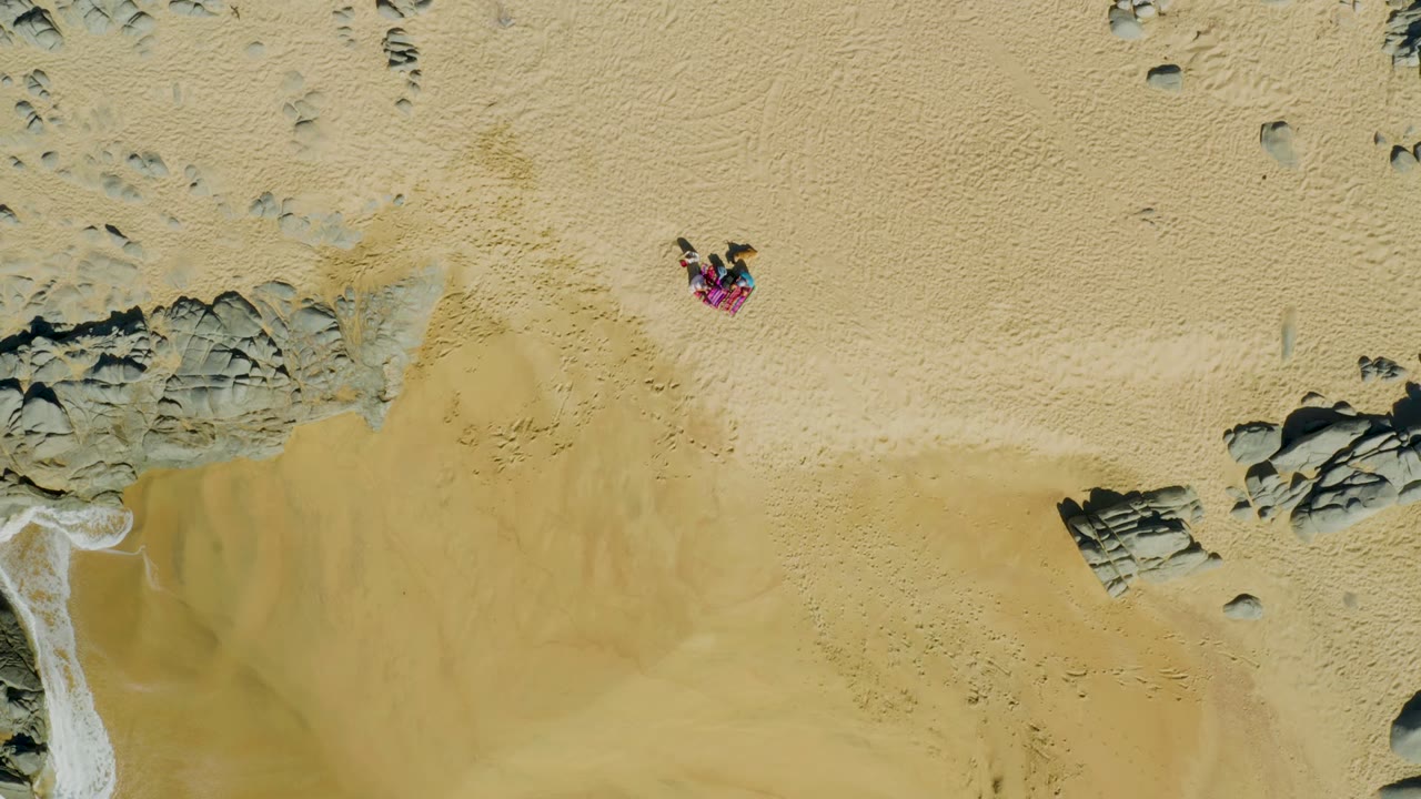 Person on a towel on pale sand with rocky shoreline and wet sand below.