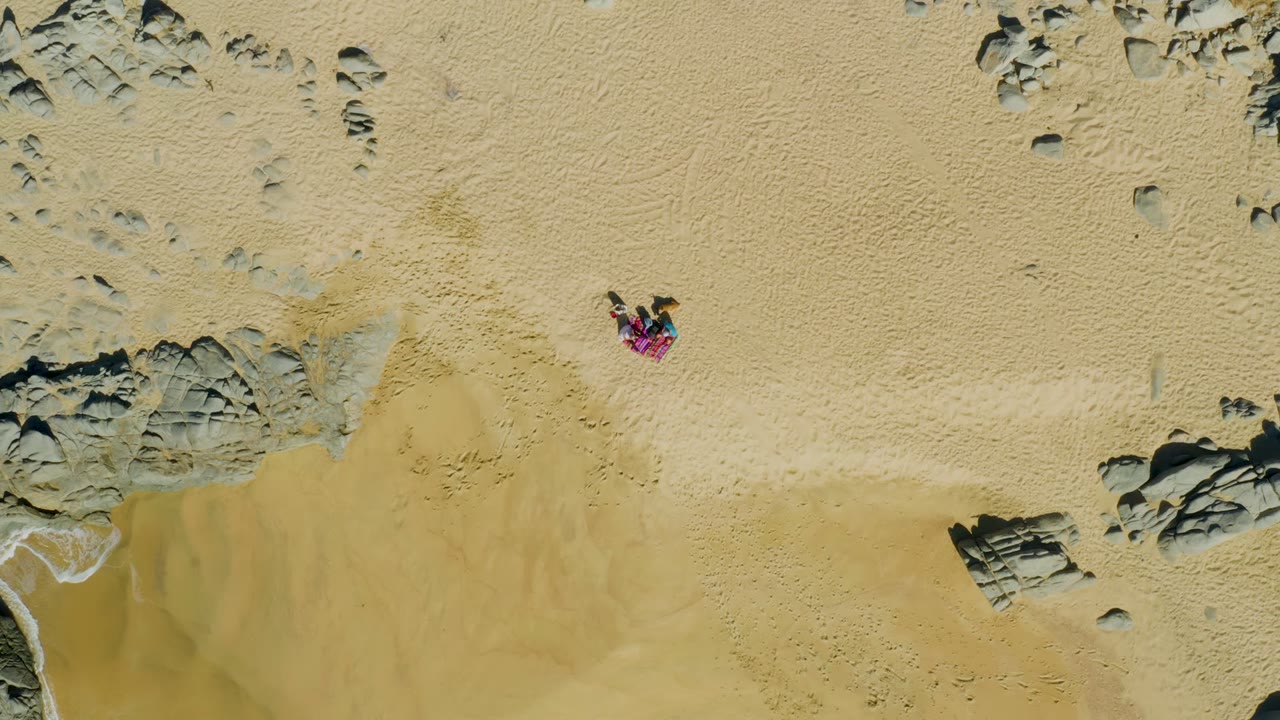 The overhead La Fortuna beach frame shows the towel and person near the upper sand, with darker rock shapes and exposed wet sand below as the waterline sits lower in frame.