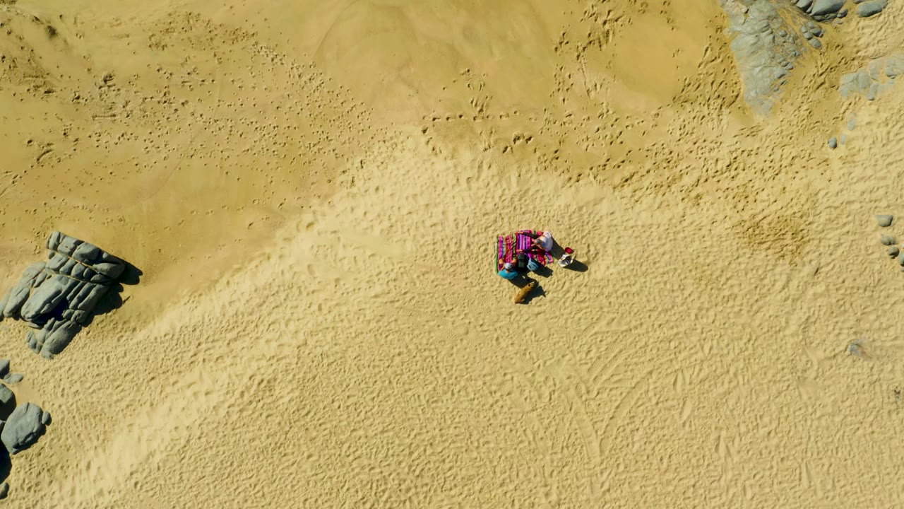 Overhead frame from La Fortuna, Baja California Sur showing a person lying on a pink towel on yellow sand, with dark rocks entering from the left edge.