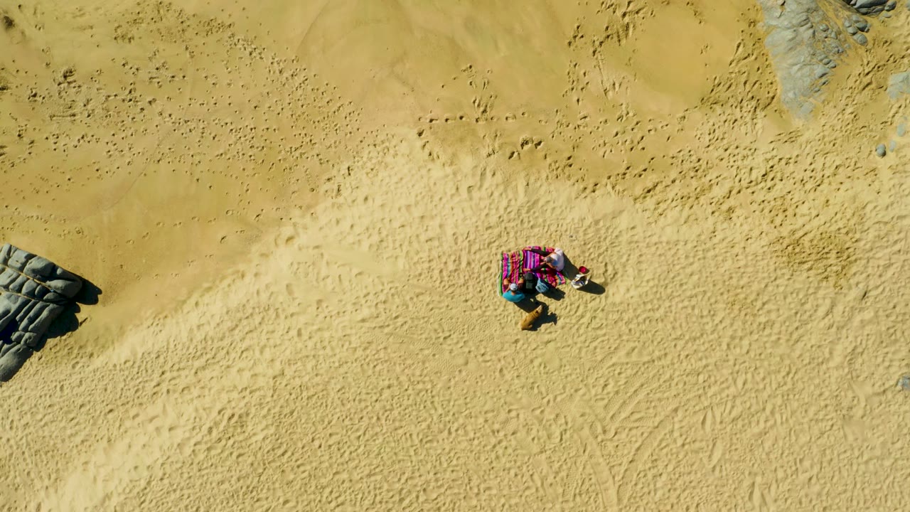 At 0:02, the overhead La Fortuna beach shot places the person on the pink towel in the right half of the frame, with dark rocks near the lower left and upper right edges.