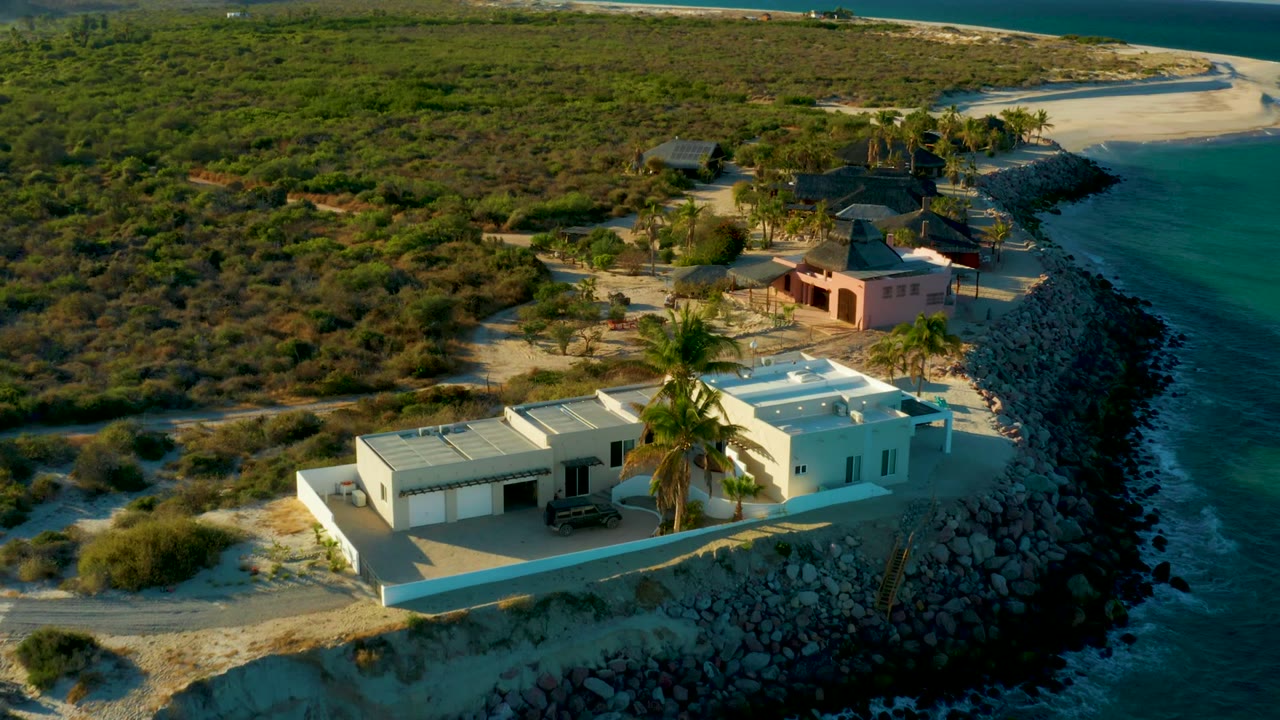 Elevated view in Las Barracas, Baja California Sur showing a white flat-roof house, palm tree, rocky shoreline, blue water, sandy ground, and no visible people.