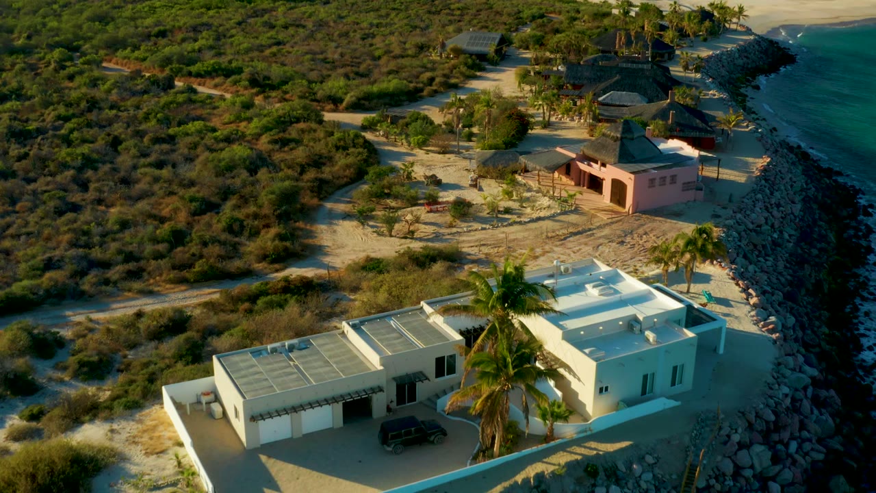 High angled drone view in Las Barracas showing the white house at lower right, the pink building and thatched roof structures beyond, a narrow rocky shoreline, blue-green water, and no visible people.