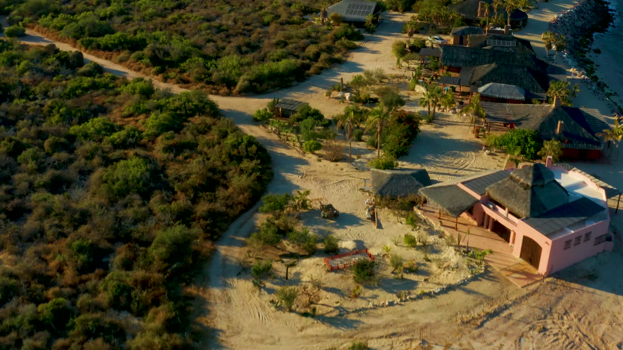 Opening aerial frame over Las Barracas shows a winding pale dirt road, dense scrub, palm trees, thatched roofs, and a pink building, with no visible people.