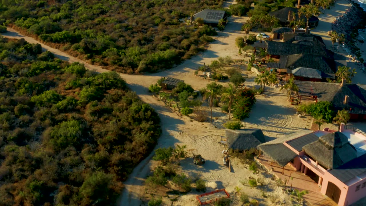 Drone view over a curved dirt track and huts surrounded by bushes.
