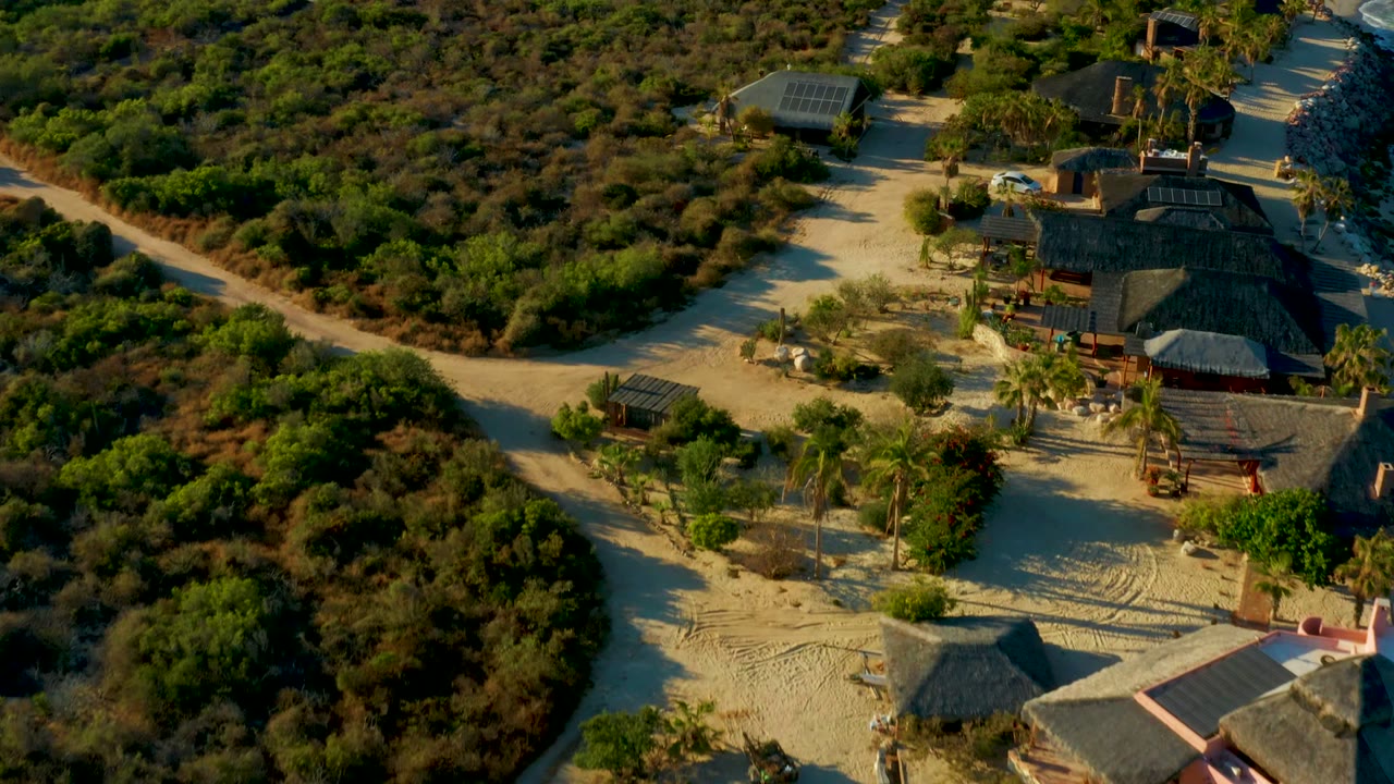 At two seconds, the drone view shows more of the thatched building cluster beside the pale road, with a roofed structure in the upper area and no visible people.