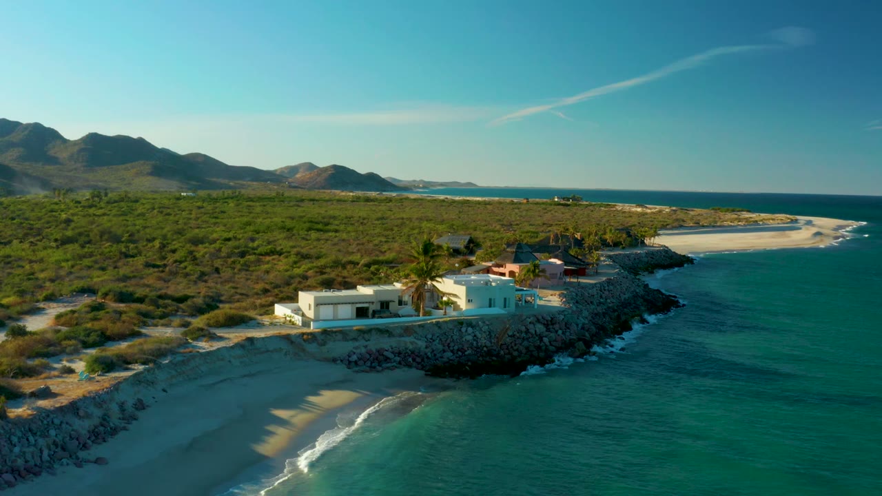 Opening aerial frame over Las Barracas shows a white house on a rocky point, turquoise water in the foreground, sandy beach to the left, green brush and hills behind, and no visible people.