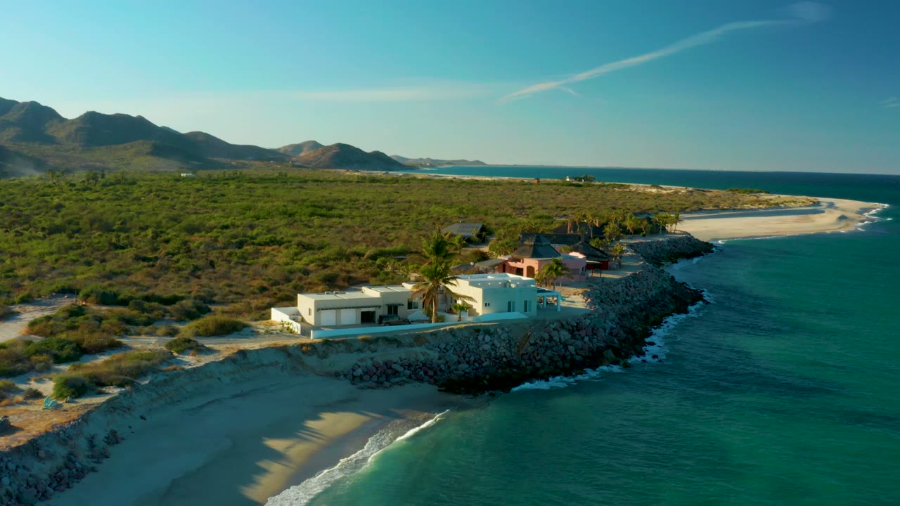Drone view of beach houses on a rocky shoreline with hills behind.