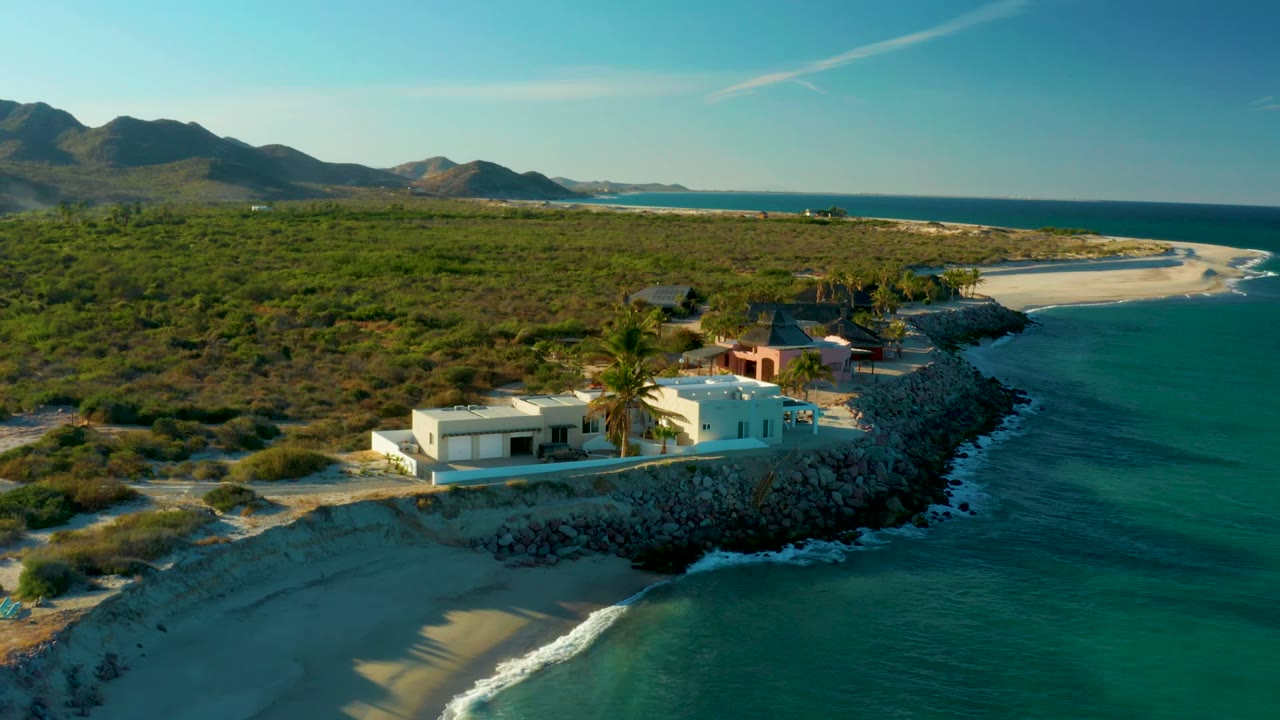 At 0:02, the drone angle places the ocean along the right edge, the white house and palms near the lower center, the pink structure behind them, and no visible people in Las Barracas.
