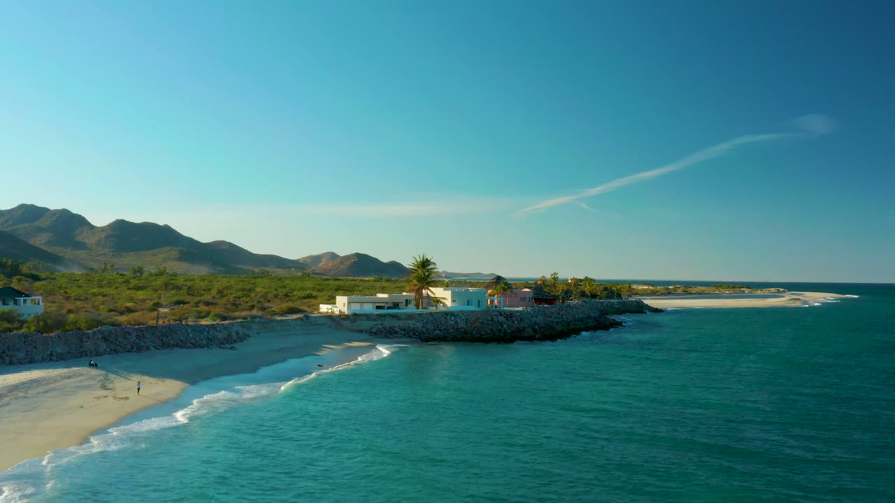 Opening aerial frame over Las Barracas, Baja California Sur, with turquoise water at right, a sandy beach at left, tiny walkers near the shore, a white beachfront building, palm trees, rocky shoreline, and mountains under clear sky.