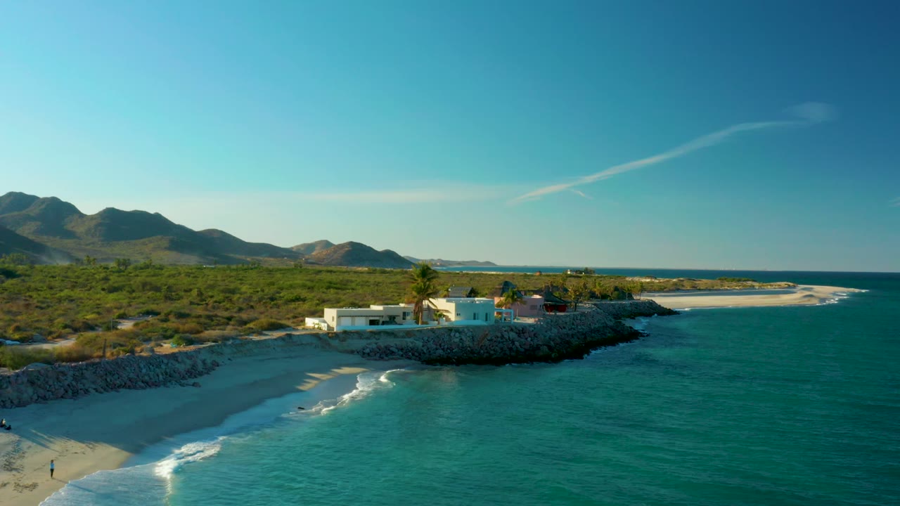 Las Barracas aerial frame with a wider sweep of green scrubland behind the beach, gentle waves reaching the sand, tiny people near the lower left beach area, a white coastal building and palms, and mountains across the horizon.