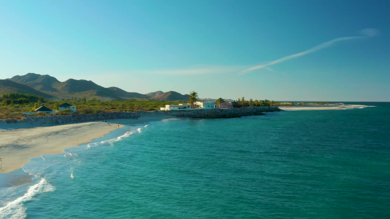 Aerial frame over Las Barracas showing turquoise water, a sandy beach at left, low coastal buildings, palms, scrubland, distant mountains, and a tiny walker near the surf.