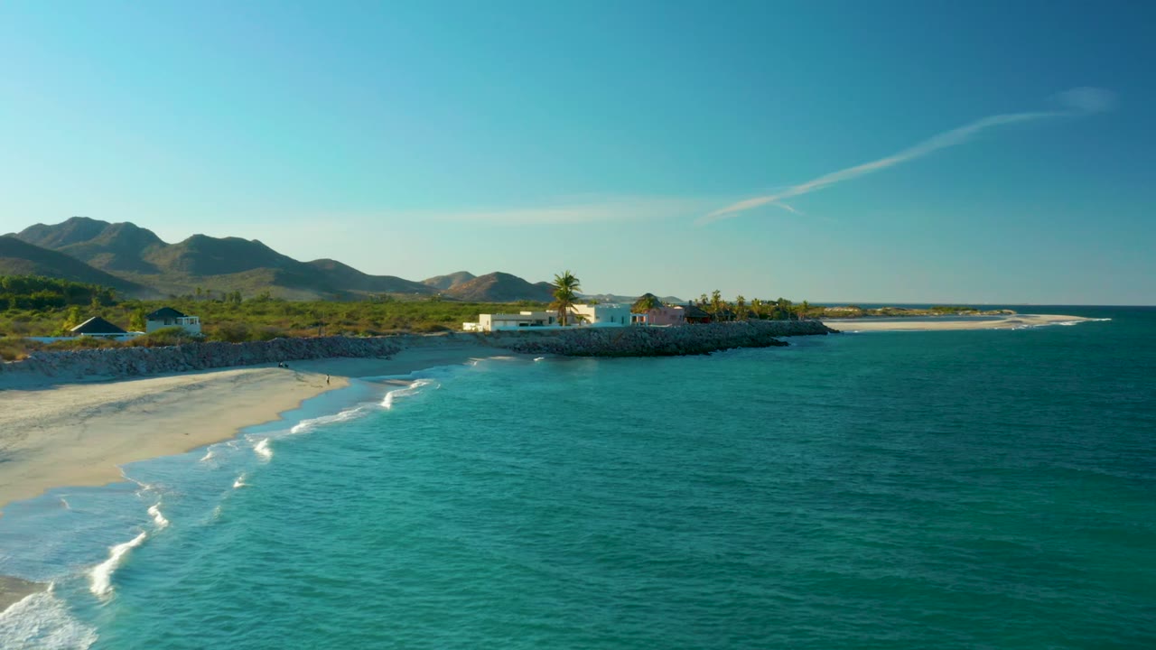 Turquoise waves roll toward a sandy beach below mountains and coastal buildings.