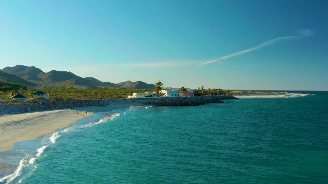 Las Barracas coastline from above, with the beach curving in from the left, turquoise water occupying the center and right, a few low buildings and palms on shore, and dark mountains in the background.