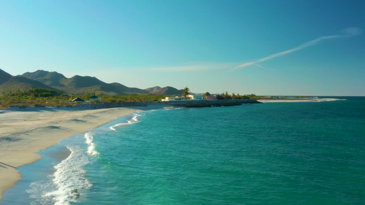 Aerial view over Las Barracas, Baja California Sur, with turquoise water on the right, pale sand on the left, distant mountains, sparse shoreline buildings, and no visible people.