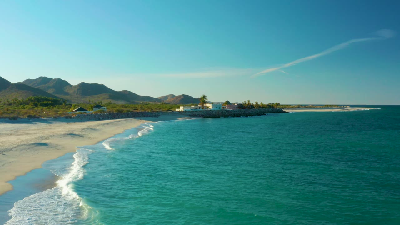 Small waves break along a sandy shoreline beside clear turquoise water and distant mountains.