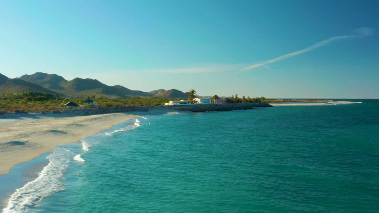 At 0:02, the Las Barracas shoreline remains wide in frame, with white foam tracing the beach at left, open turquoise sea at right, distant buildings, mountains, and no visible people.