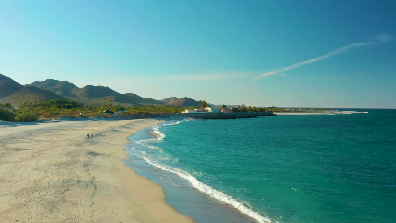 Aerial frame from Las Barracas, Baja California Sur, showing turquoise ocean on the right, pale sand on the left, two distant beach walkers, low coastal buildings, and hills under a clear blue sky.