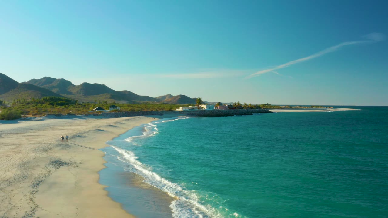 Wide aerial frame over Las Barracas with turquoise sea occupying the right half, foam bands rolling onto pale sand, two distant people on the beach, low buildings, and hills along the horizon.