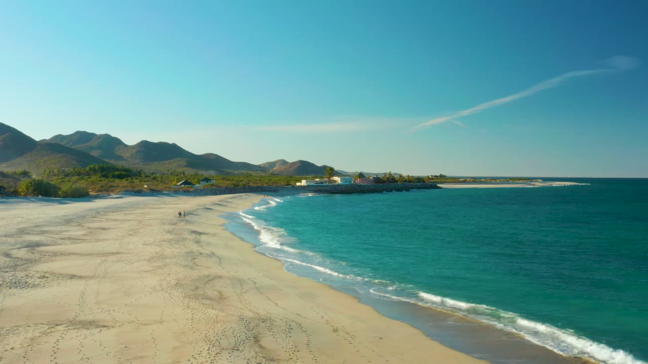 Opening frame of Las Barracas, Baja California Sur, showing a broad sandy beach, turquoise surf on the right, distant mountains, coastal buildings, and tiny walkers on the sand.