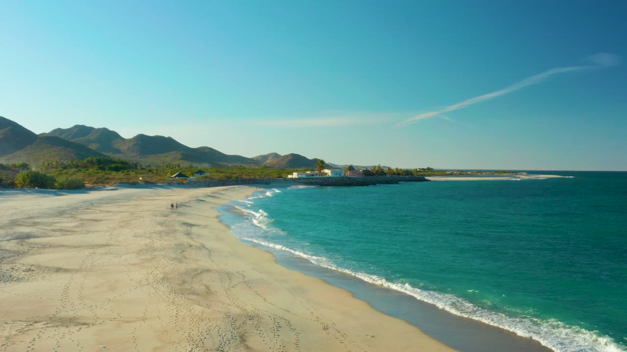 Las Barracas coastline at 0:02 with the beach sweeping from left foreground toward the distant buildings, turquoise water occupying the right half, and small walkers remaining visible on the sand.
