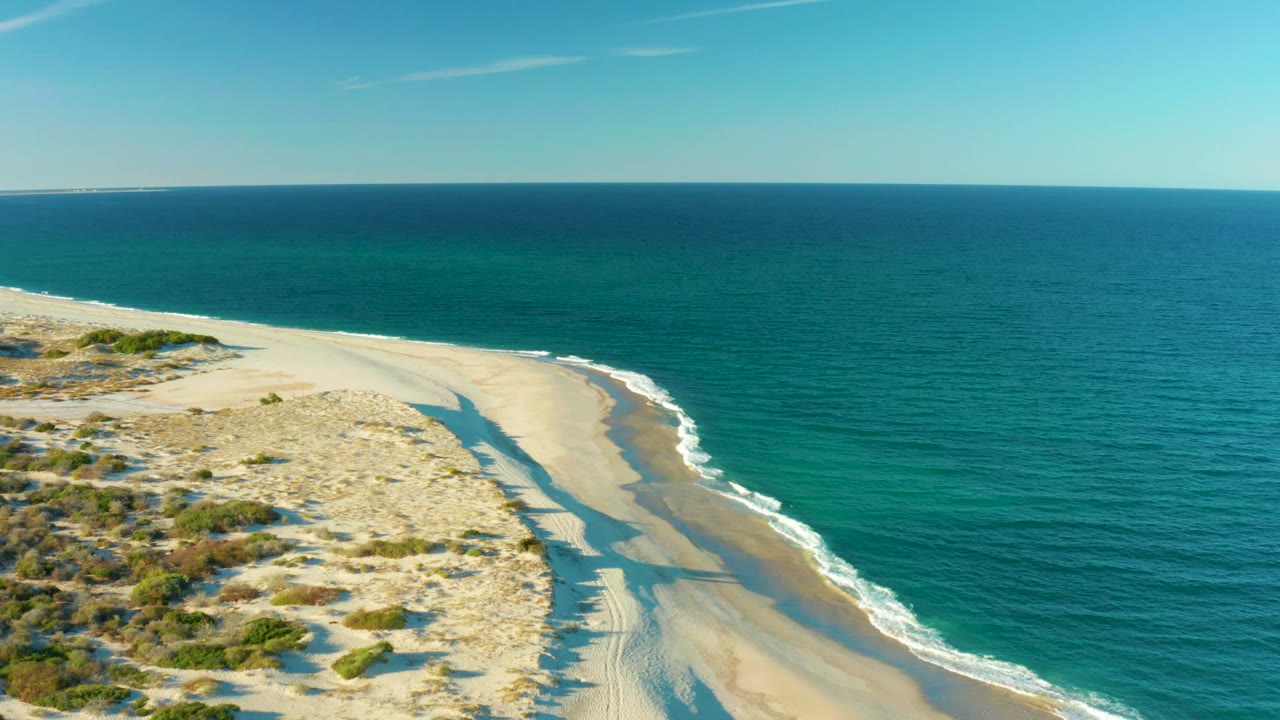 Aerial frame over Las Barracas, Baja California Sur, with sandy dunes at left, a curving beach, turquoise ocean at right, and no visible people, though a long shadow crosses the sand.