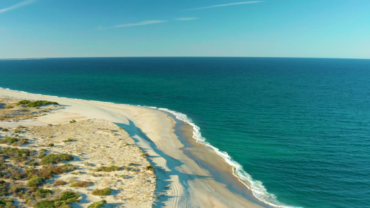 Drone view of pale beach dunes beside calm turquoise ocean.