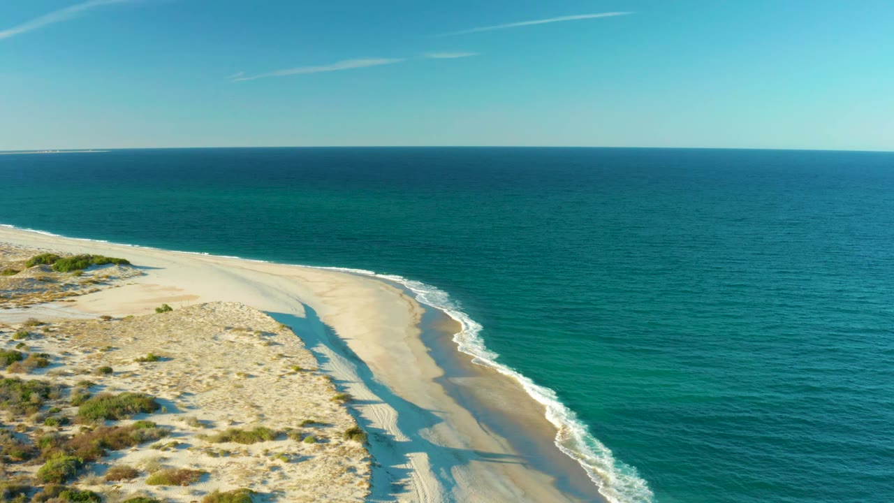 Aerial frame over Las Barracas showing textured dunes in the lower left, a bright sand beach, a thin white surf edge, open turquoise ocean, and no visible person besides the shadow on sand.