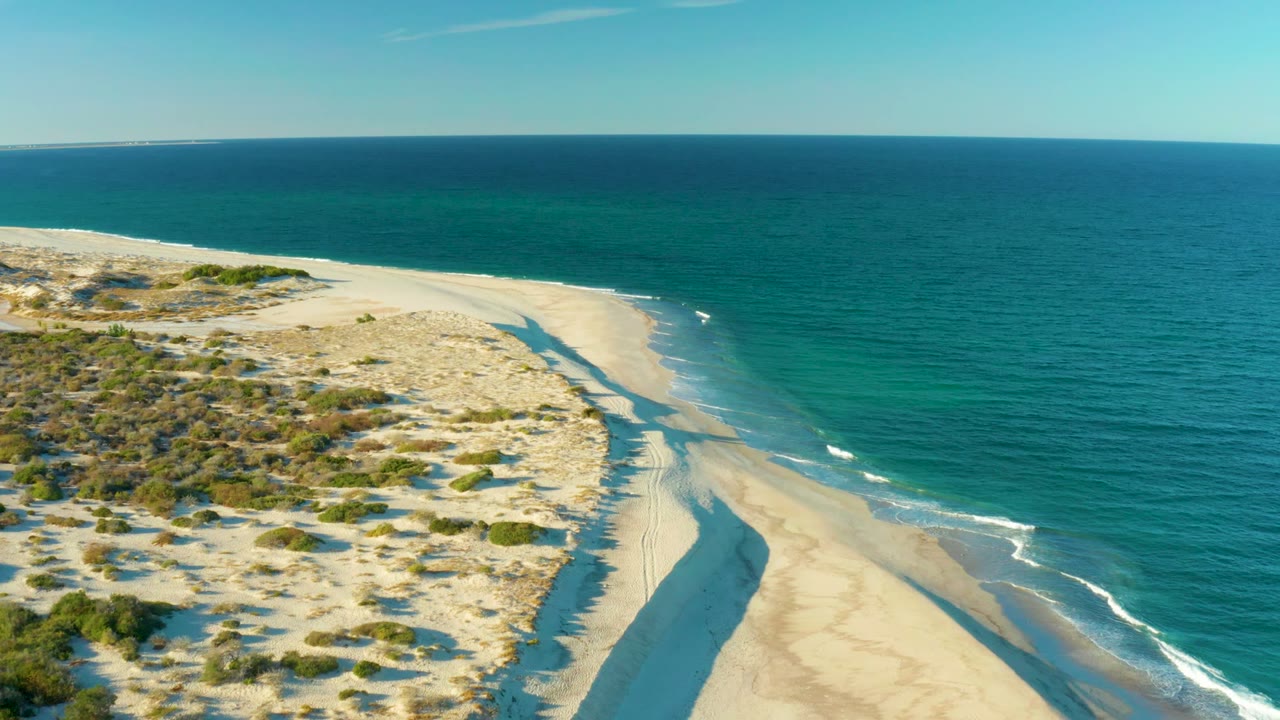 Drone view of dune-covered sand and gentle surf along the shore.