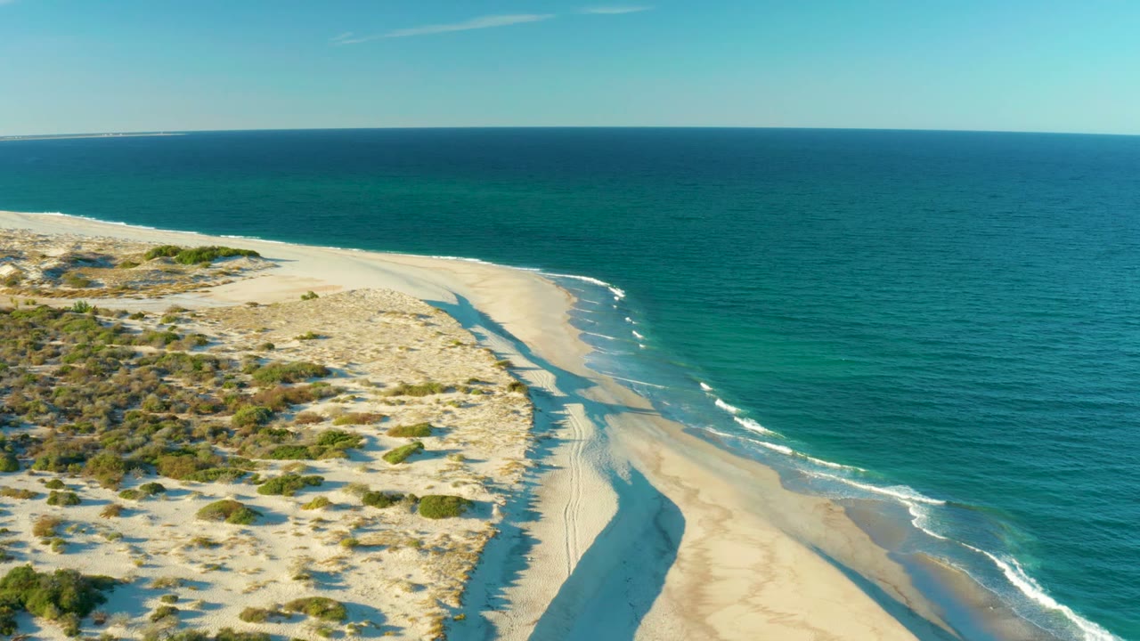 High aerial view of Las Barracas with sunlit dunes at left, a curved strip of wet sand, small white wave breaks, deep blue ocean, and no visible people.