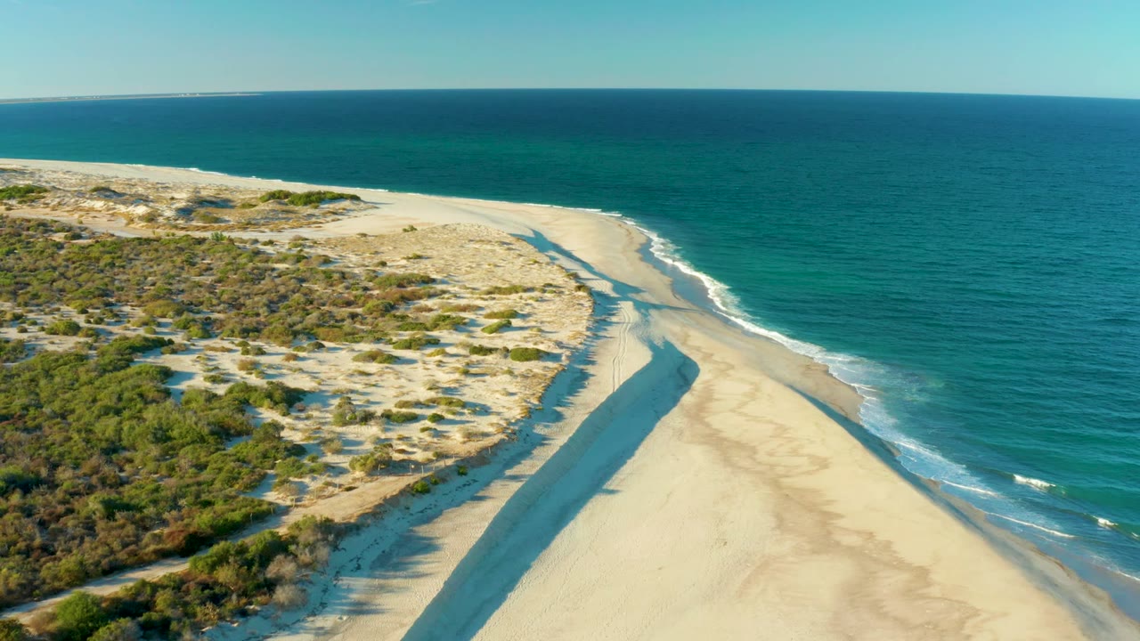 Aerial frame over Las Barracas showing a sandy spit bordered by low green dunes and turquoise-to-blue ocean, with no visible people.