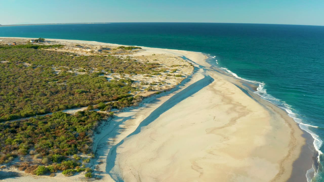 Aerial view over Las Barracas showing pale sand, a narrow tidal channel, green dune vegetation, turquoise ocean, and no visible people at the opening moment.