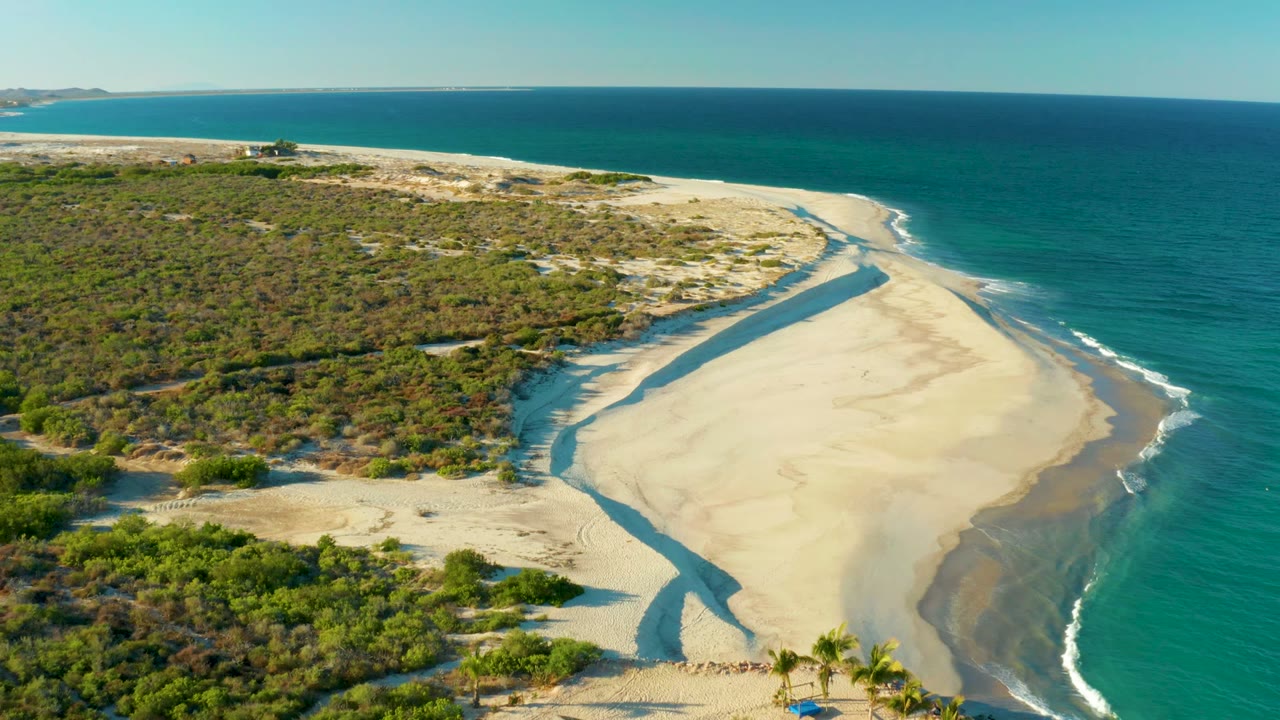 Aerial frame over Las Barracas, Baja California Sur, showing scrub-covered dunes, a pale sand spit, turquoise ocean, a dark channel across the beach, small beach structure near the lower edge, and no visible people.
