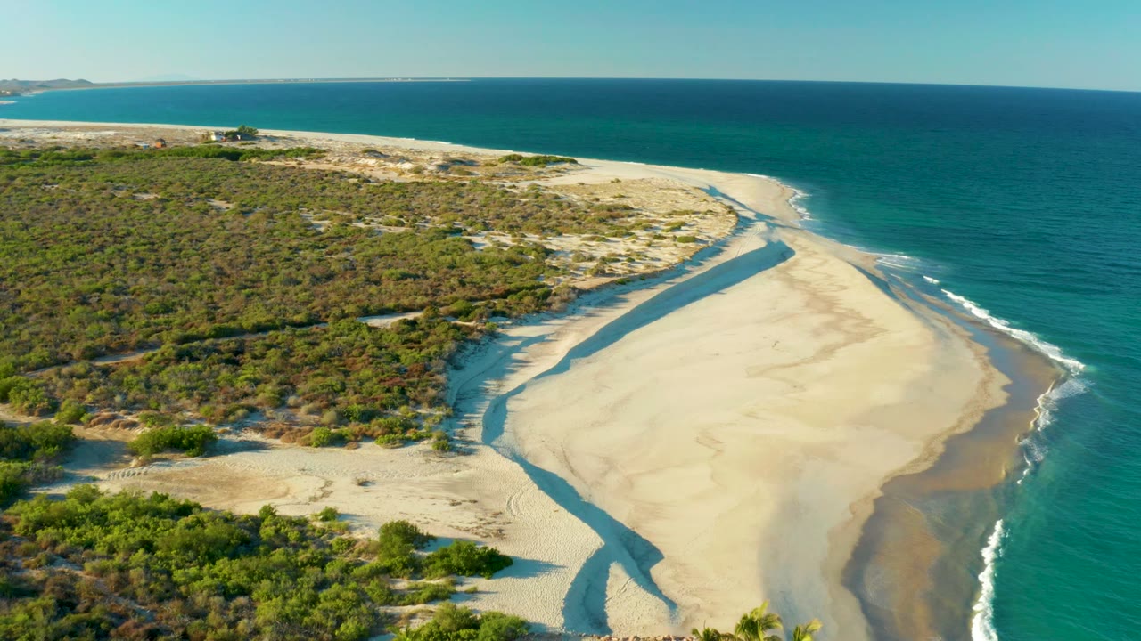 Drone view of dunes and a sandy shoreline with waves beside a narrow channel.
