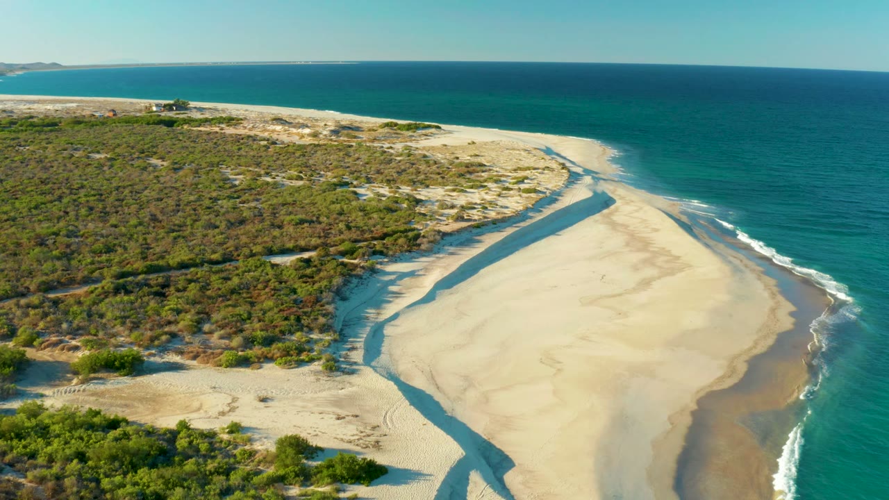Aerial frame over Las Barracas showing the beach channel angled through the sand, scrubby dunes to the left, turquoise sea to the right, white foam along the shoreline, distant land on the horizon, and no visible people.