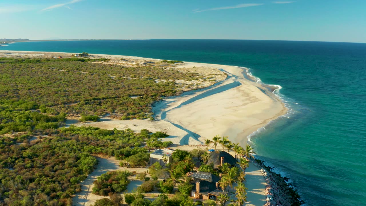 Aerial frame over Las Barracas, Baja California Sur, Mexico, showing a sandy spit curving beside turquoise water, scrub vegetation inland, palms and buildings in the foreground, and tiny distant people near the shore.