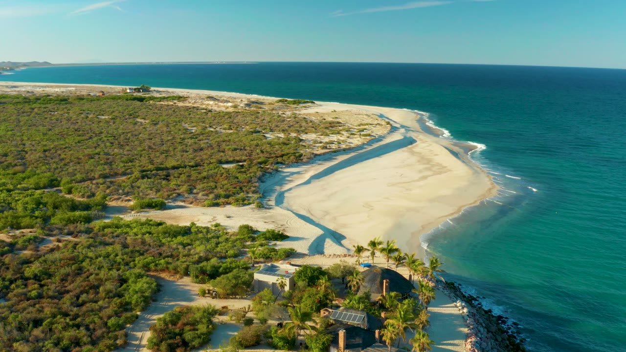 Wide drone view of sandbar, narrow water channel, ocean waves, brush, palms, and buildings.