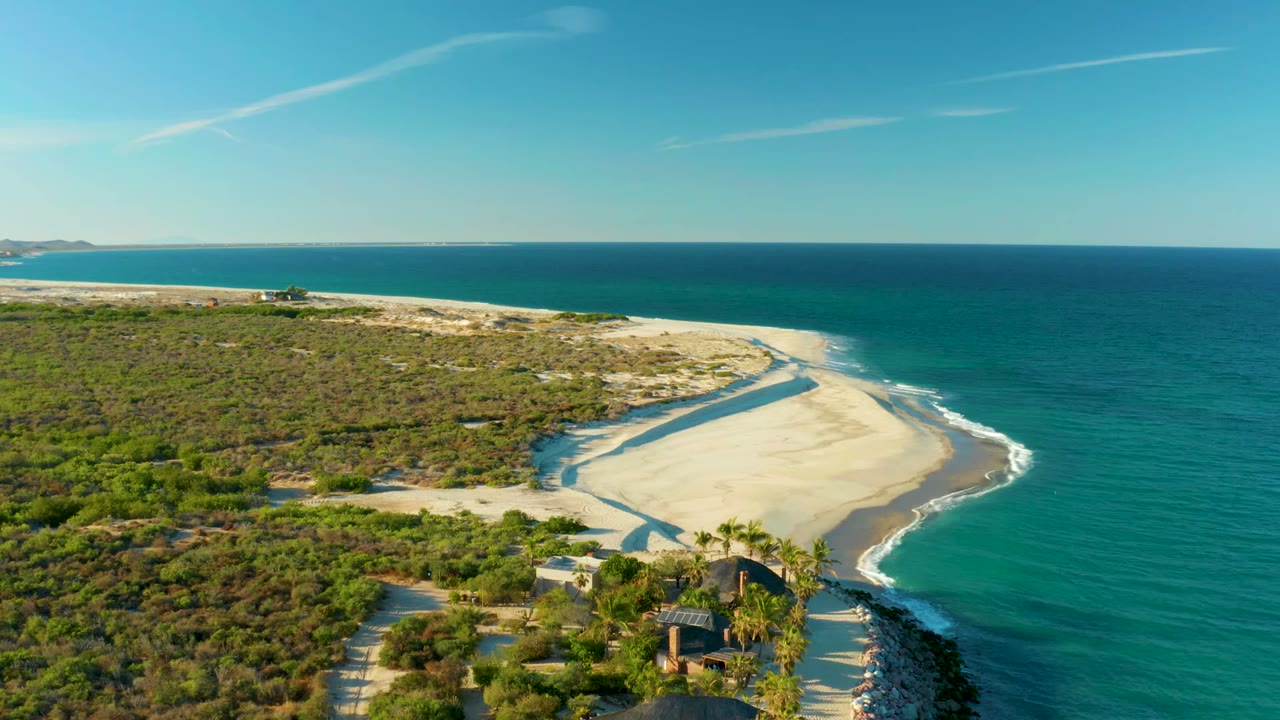 Aerial frame over Las Barracas showing a curved pale sand beach bordered by turquoise ocean and scrub vegetation, with small structures near palms in the lower frame and no visible people.