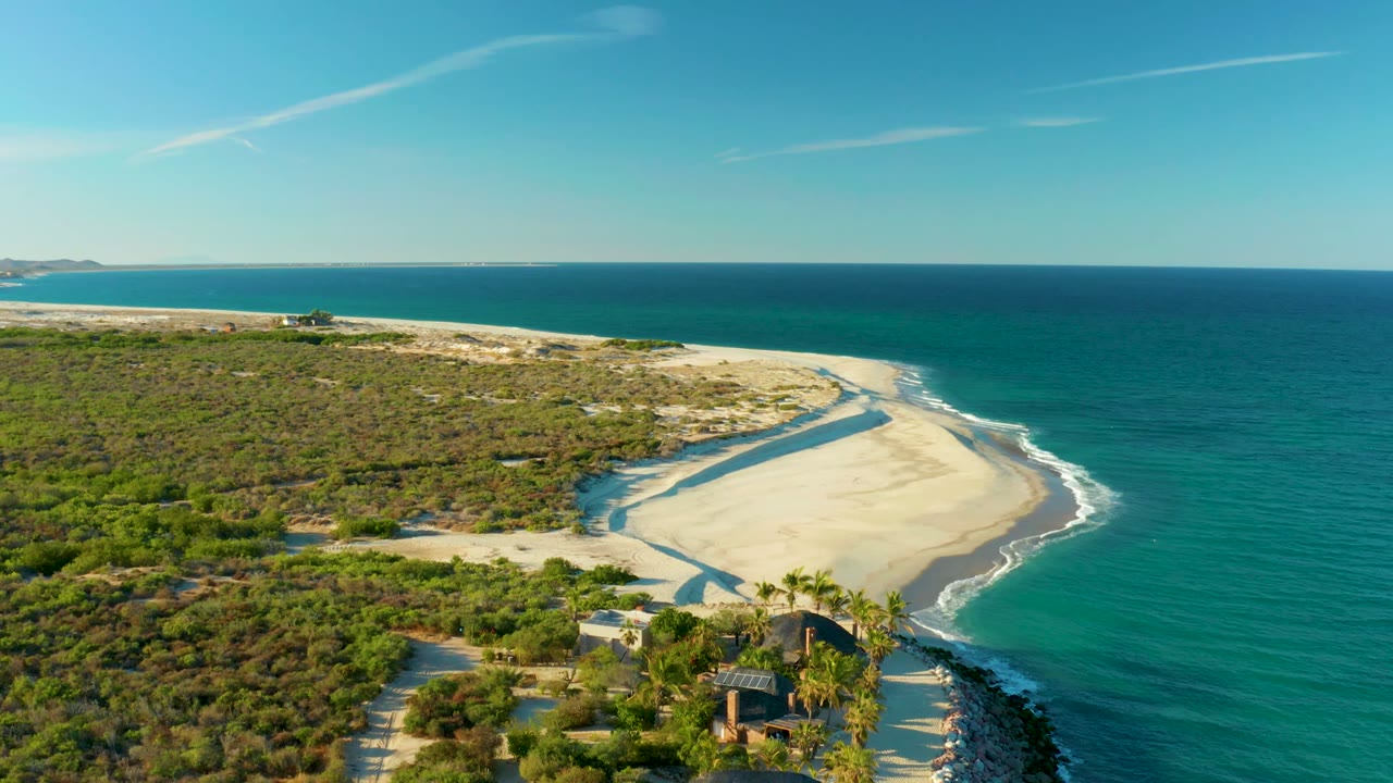 High aerial frame of the Las Barracas shore with pale sand curving toward the right, turquoise water beyond, low vegetation across the left side, distant coastline on the horizon, and no visible people.