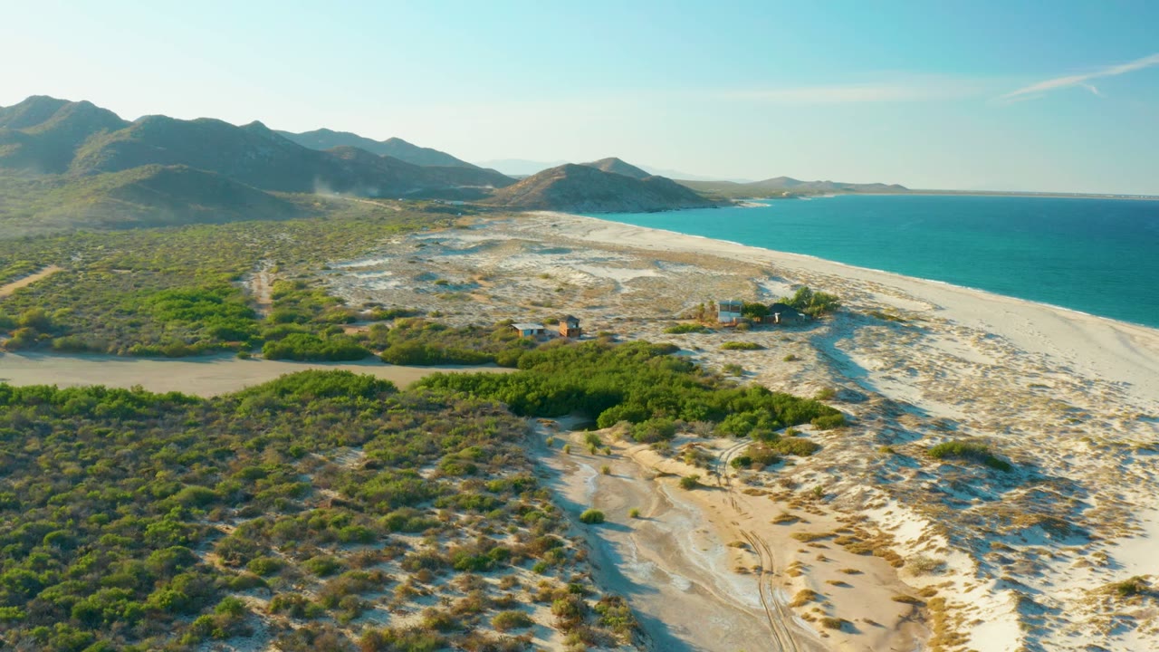 Aerial view over Las Barracas, Baja California Sur, with a sandy river channel leading toward turquoise ocean, green scrub in the foreground, mountains in the distance, and no visible people.