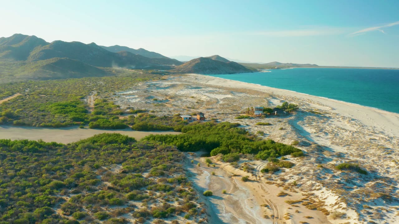 Coastal aerial frame over Las Barracas with scrubland and sand tracks in front, a muted river inlet crossing the midground, bright beach and ocean to the right, mountains beyond, and no visible people.