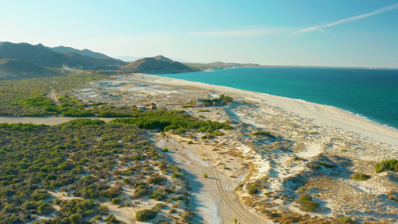 Aerial frame over Las Barracas, Baja California Sur showing a sandy beach beside blue ocean, a dirt path through coastal scrub, distant mountains, and no visible people.