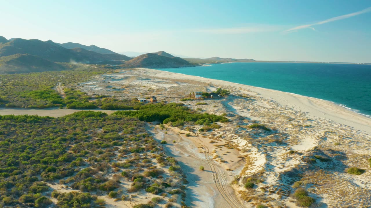 Drone view of coastal scrub, sandy track, beach, ocean, and mountain horizon.