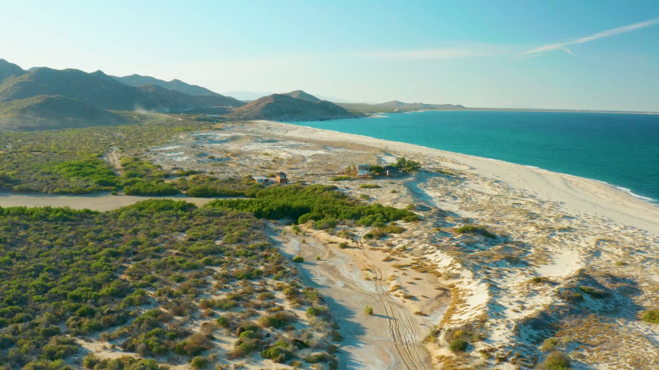 Wide aerial frame over the Las Barracas coastline showing a sandy shore curving along blue water, scrubby terrain and dirt tracks inland, distant structures, mountains, and no visible people.