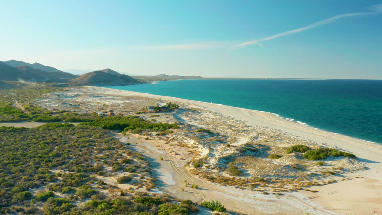 Aerial frame from Las Barracas, Baja California Sur, showing sandy dunes in front, turquoise ocean along the right edge, scrub vegetation to the left, distant mountains, and no visible people.