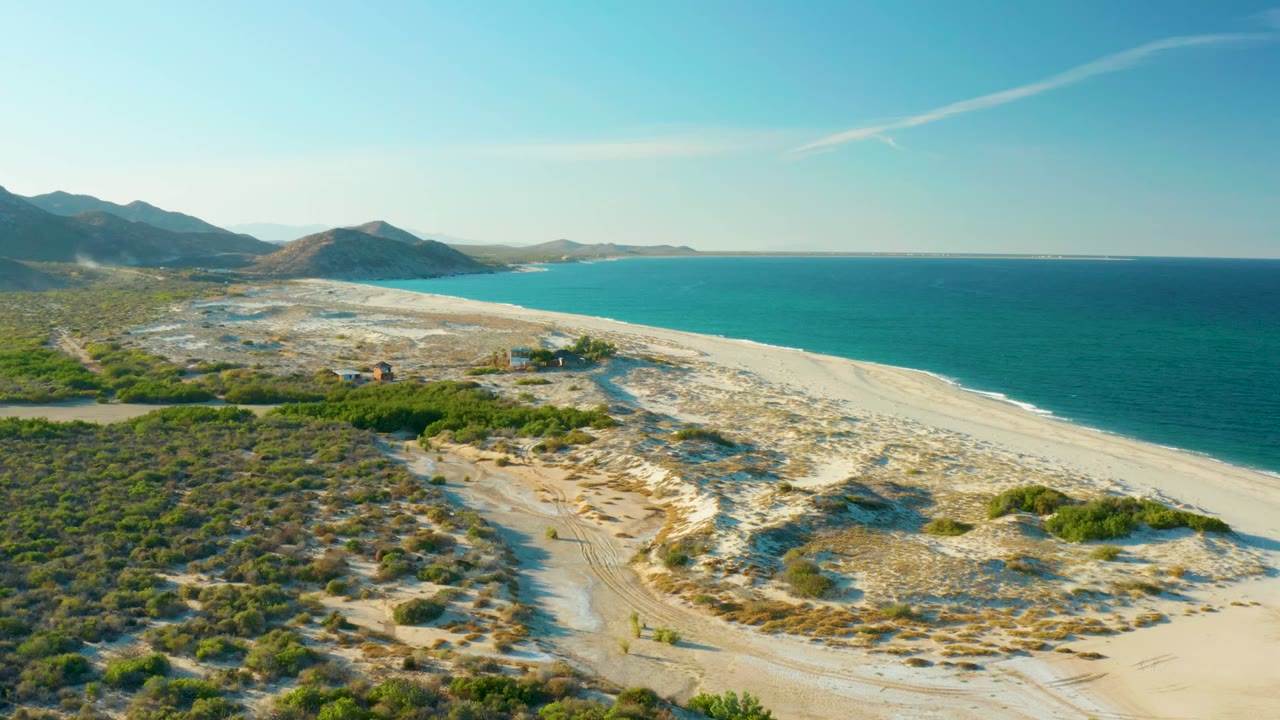 Second aerial frame over Las Barracas coastline, with pale dunes and track marks below, blue-green ocean on the right, low mountains on the horizon, and no visible people.